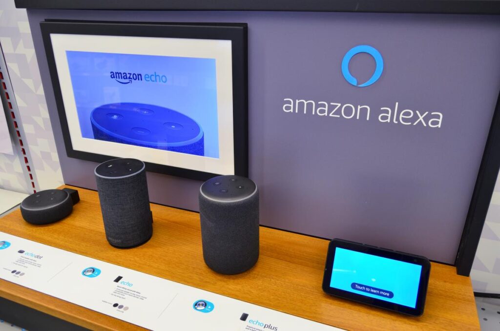 A desk with a collection of Amazon Alexa products on it, with the Amazon Alexa logo on the wall behind it