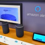 A desk with a collection of Amazon Alexa products on it, with the Amazon Alexa logo on the wall behind it