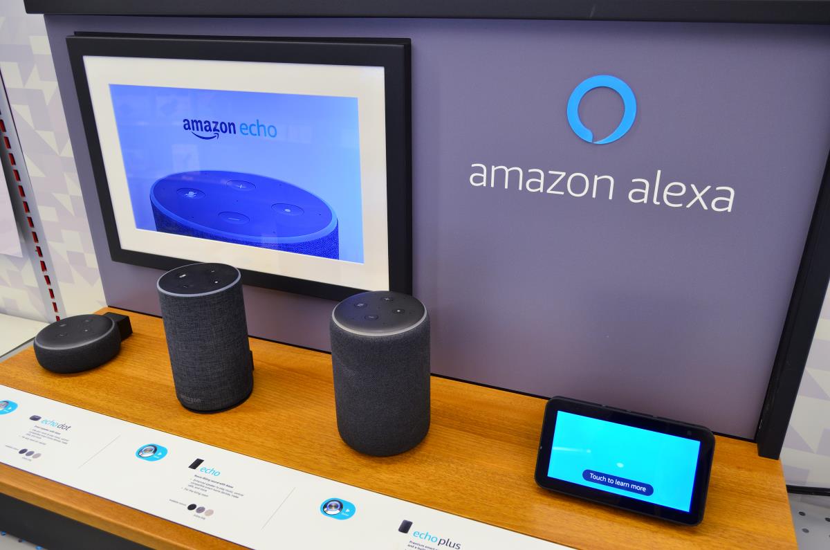 A desk with a collection of Amazon Alexa products on it, with the Amazon Alexa logo on the wall behind it