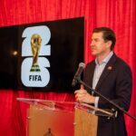 A man standing at a podium next to a TV, and on the TV is the branding for the 2026 FIFA World Cup