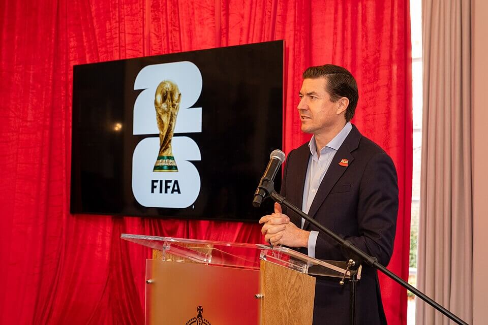 A man standing at a podium next to a TV, and on the TV is the branding for the 2026 FIFA World Cup