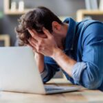 A man sitting at a table with his laptop, and has his head in his hands, looking frustrated