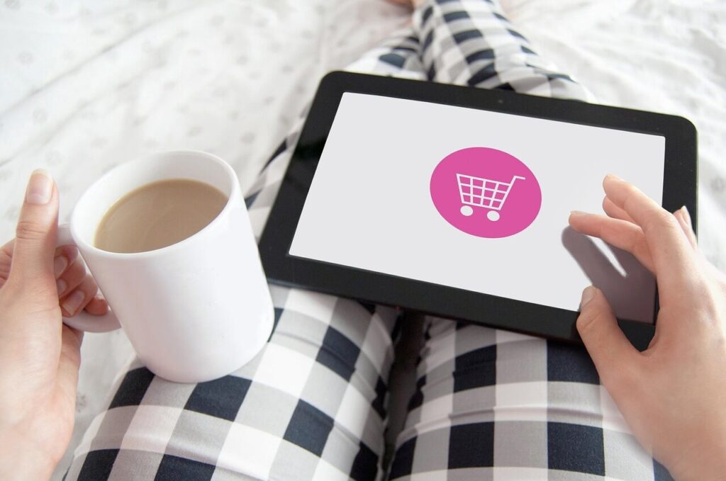 A person sitting on a couch with their coffee in one hand, and a tablet sitting on their lap, and it has a shopping cart on the screen.