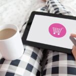 A person sitting on a couch with their coffee in one hand, and a tablet sitting on their lap, and it has a shopping cart on the screen.