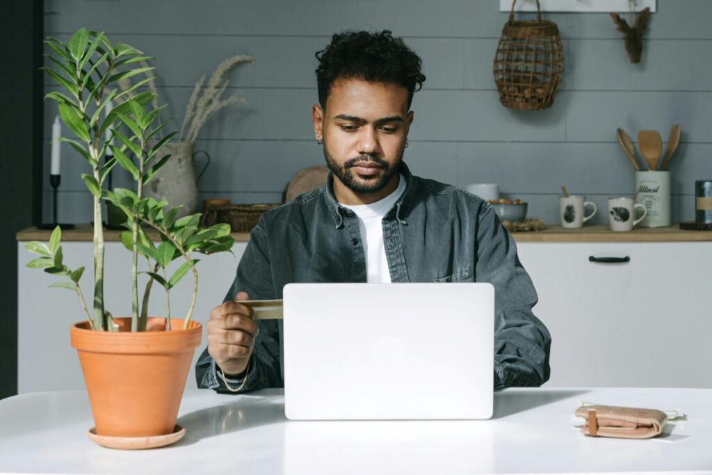 A man staring at his computer, typing, and also holding a credit or debit card in his hand
