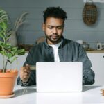 A man staring at his computer, typing, and also holding a credit or debit card in his hand