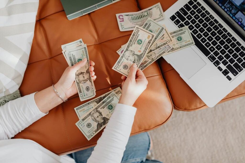 Person counting money on a couch with an open laptop beside them