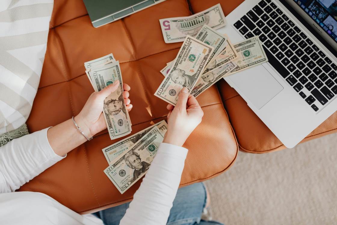 Person counting money on a couch with an open laptop beside them